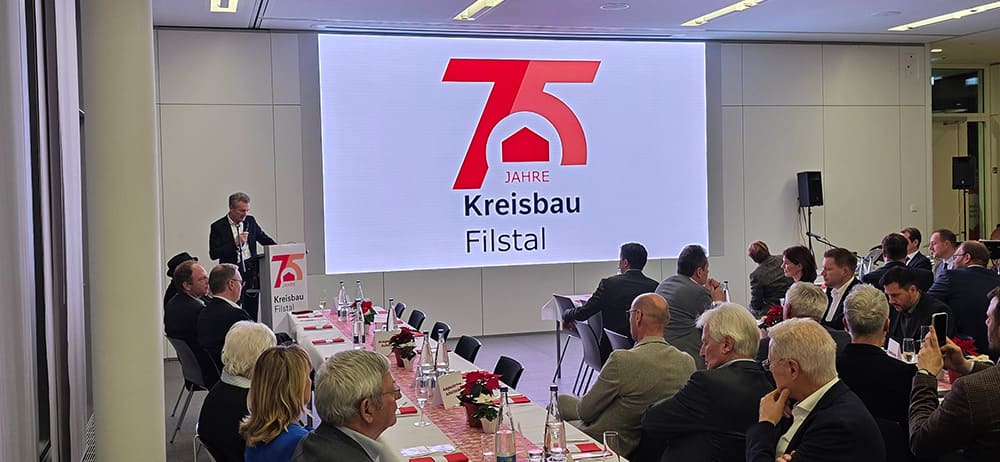 Vortrag in einem Konferenzraum mit mehreren sitzenden Personen und einem Redner am Pult vor einer großen Leinwand mit dem Logo '75 Jahre Kreisbau Filstal'
